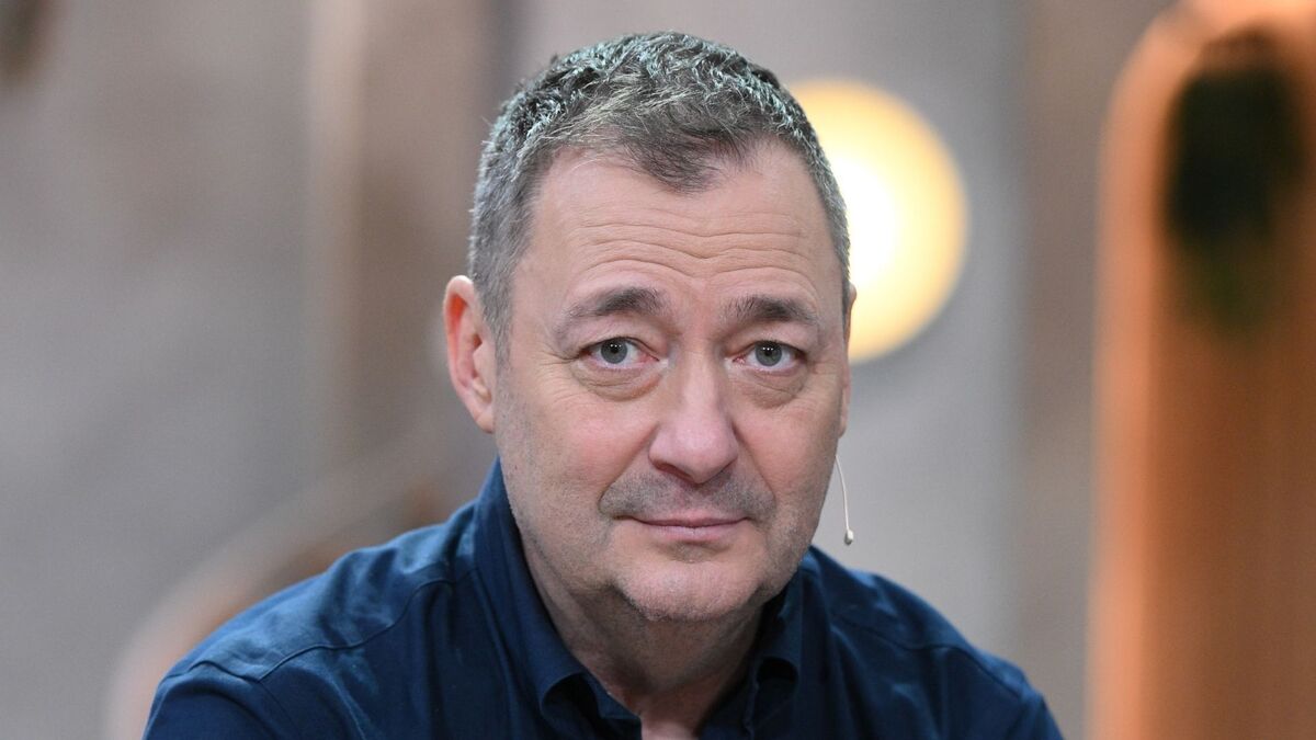 Jacek Rozenek ma krótko przystrzyżone, siwiejące włosy i jest zwrócony w stronę obiektywu. Ma na sobie ciemnogranatową koszulę oraz mikrofon nagłowny przy policzku. W tle widoczne są rozmyte elementy wnętrza i ciepłe światło.