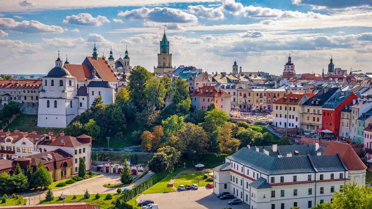 Panorama Lublina z zabytkową zabudową, kościołami i wieżami widziana z podwyższonego punktu. W centrum kadru znajduje się wzgórze z historycznymi budynkami otoczonymi drzewami. W tle widać gęstą zabudowę miasta oraz niebo z licznymi chmurami.