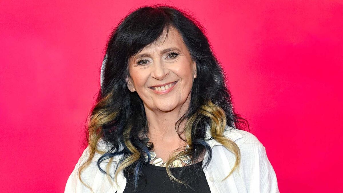 Wanda Kwietniewska ma długie ciemne włosy z jasnymi pasmami i jest ubrana w czarną bluzkę oraz białą koszulę. Na szyi ma ozdobny naszyjnik, a twarz jest skierowana w stronę aparatu. W tle znajduje się jednolita, intensywnie różowa powierzchnia.