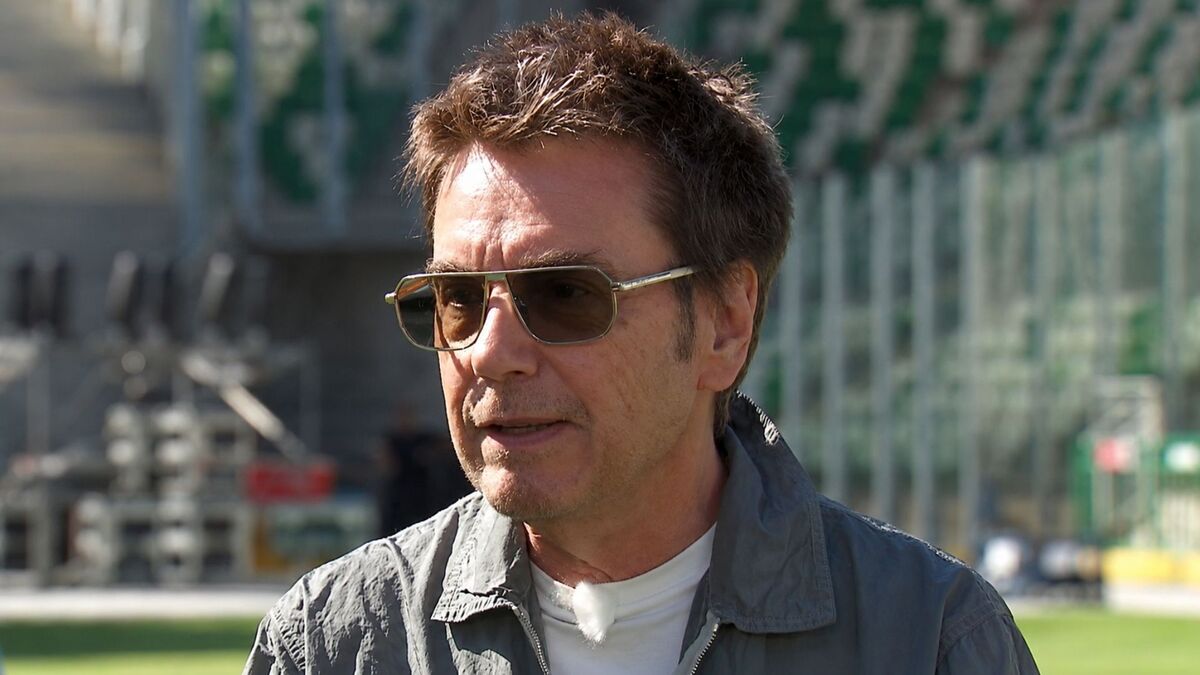 Jean-Michel Jarre podczas plenerowego wywiadu. Francuski pionier muzyki elektronicznej ma na sobie ciemne okulary przeciwsłoneczne, jasny T-shirt i szarą kurtkę. W tle widoczne są elementy infrastruktury scenicznej oraz nowoczesny budynek, a całość utrzymana jest w słonecznej, festiwalowej atmosferze.