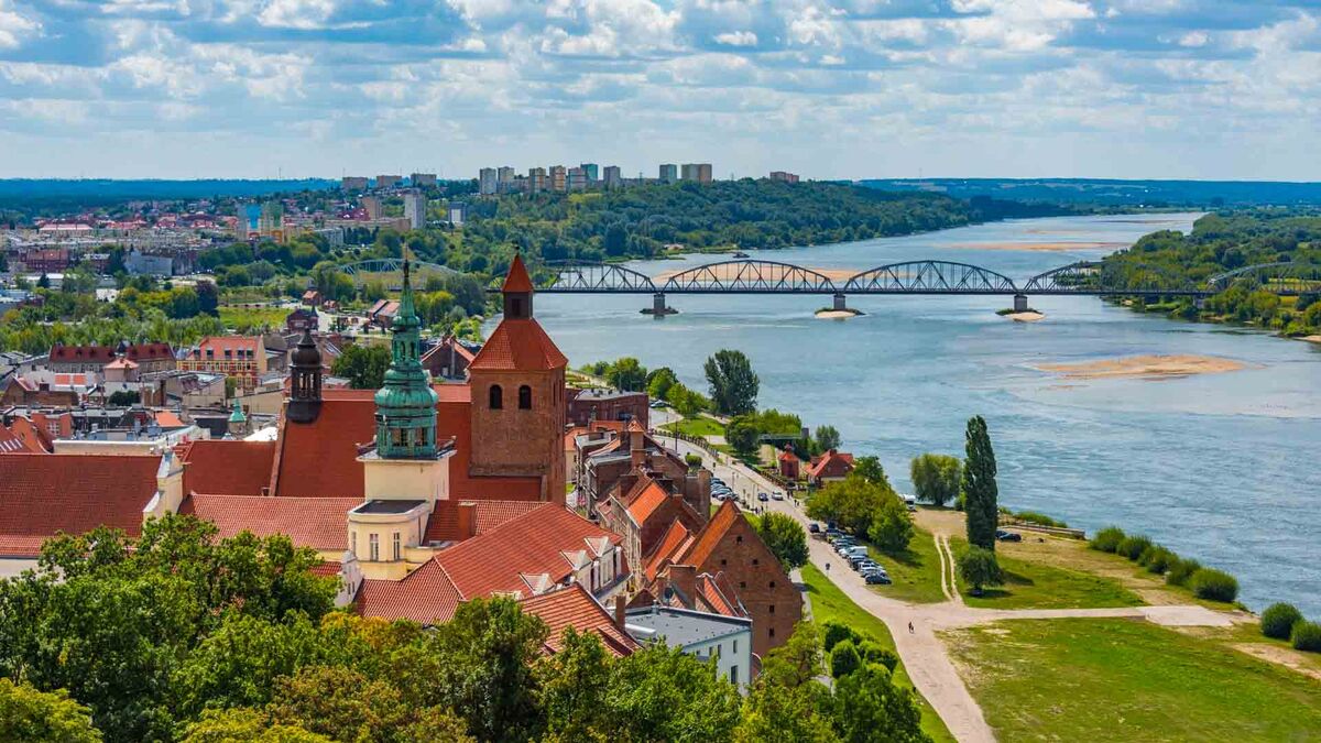 Panorama Grudziądza ukazuje zabytkową zabudowę miasta położoną nad Wisłą. Na pierwszym planie widoczne są dachy kamienic i kościołów, a w tle rozciąga się most drogowy nad rzeką. Fotografię wykonano z punktu widokowego, obejmując szeroki krajobraz miasta i doliny Wisły.